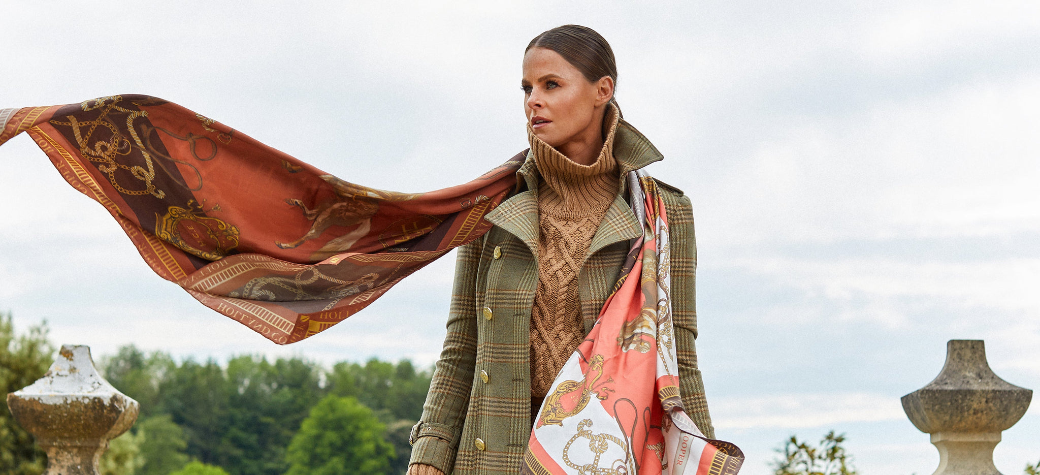 Jade Holland Cooper, owner of Holland Cooper, stood in Holland Cooper Trench Coat in a green tweed with high roll neck camel cable knit jumper and brown and orange scarf blowing in the wind