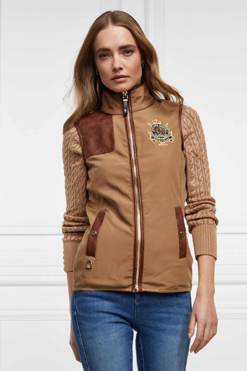 womens light brown gilet with dark brown leather seams along the arm holes pockets and down the zip with a gun patch on the shoulder and an embroidered logo worn with a beige knitted jumper and denim skinny jeans