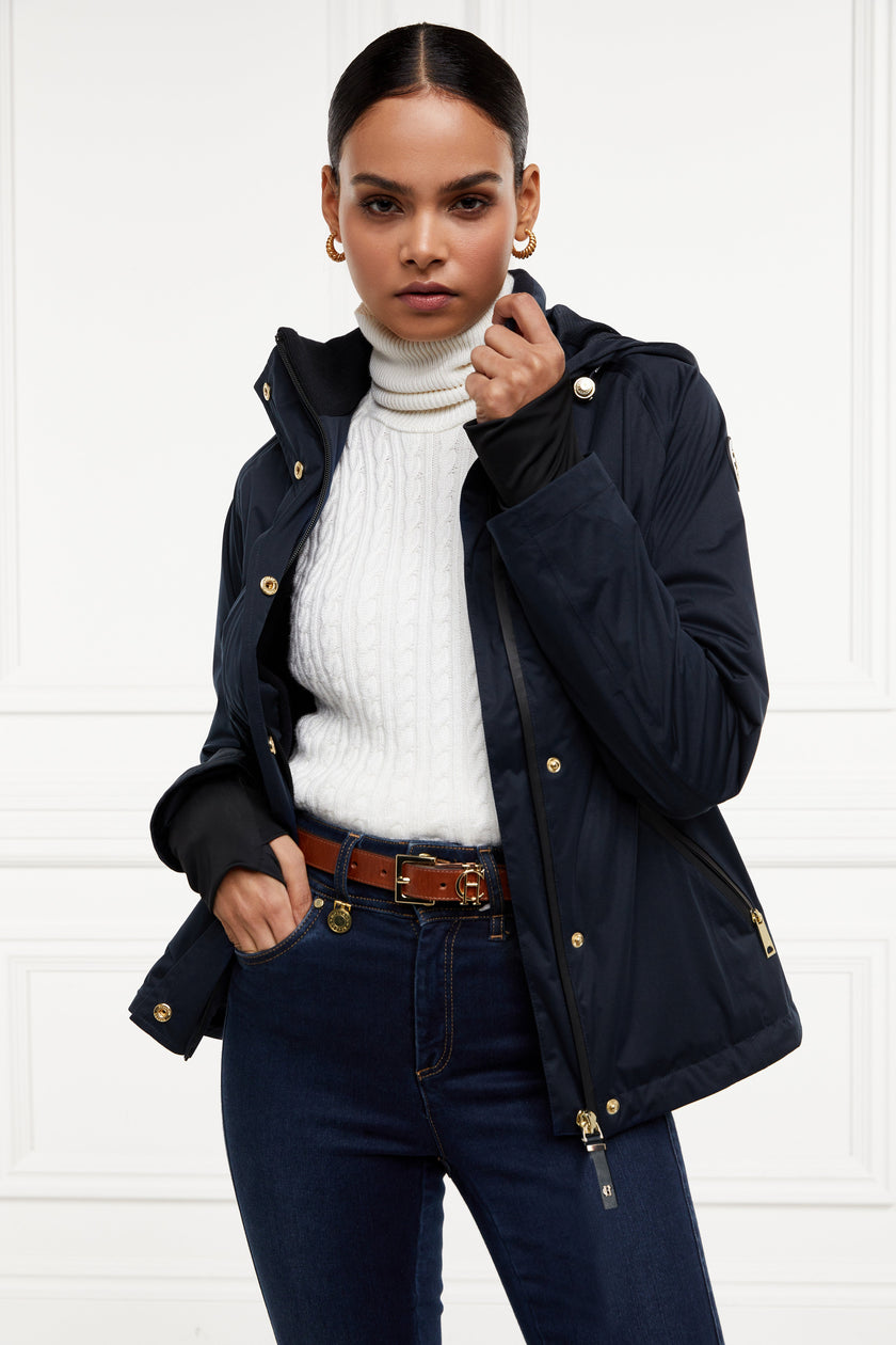 Womens waterproof navy jacket with stowaway hood storm cuffs and two pockets at hip with waterproof zip fastening styled with white cable knit roll neck indigo jeans and tan belt