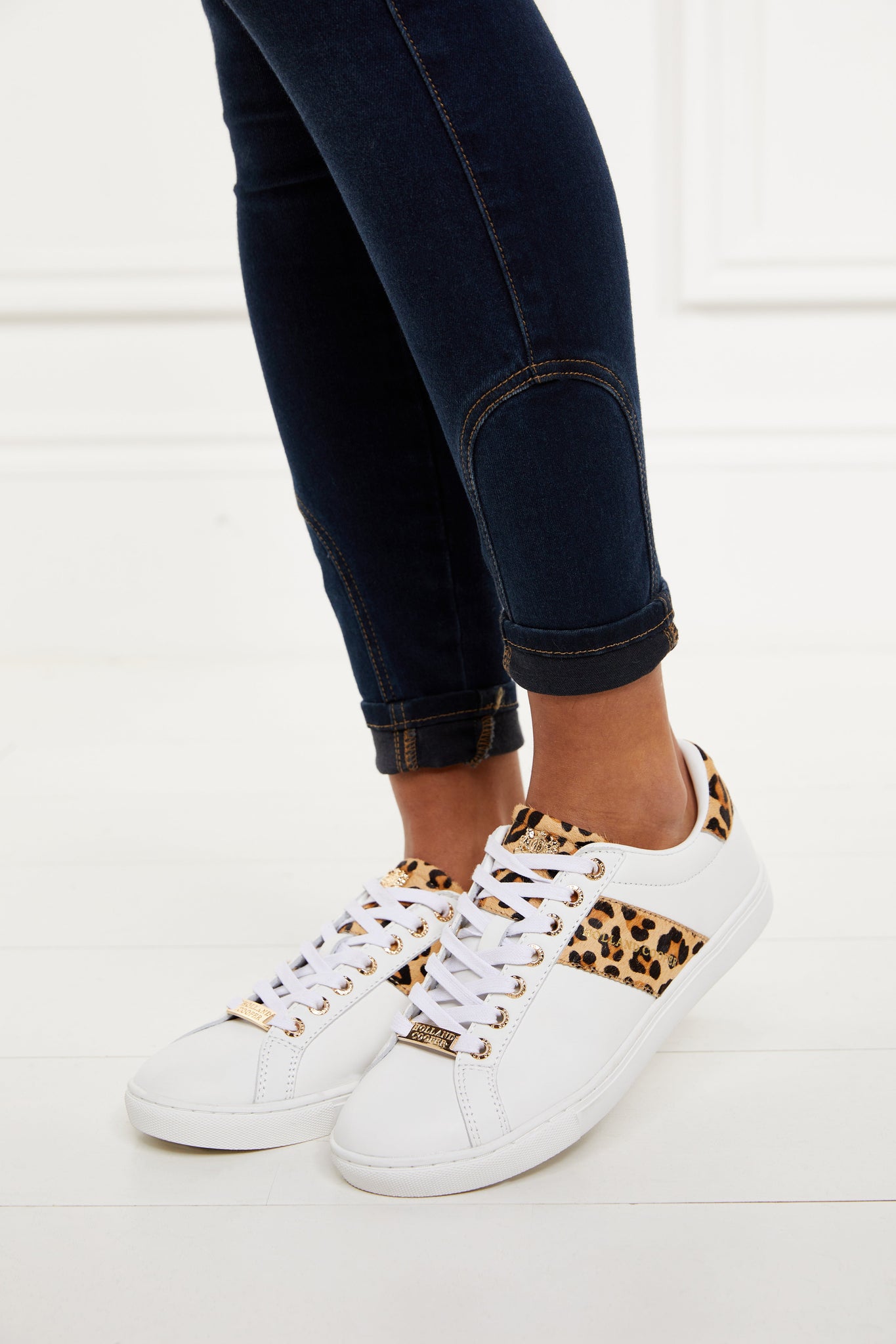 white leather trainers with white laces detailed with a diagonal stripe of leopard print on the side with gold foil branding and a leopard print heel and tongue with gold hardware. Worn with rolled up denim skinny jeans.