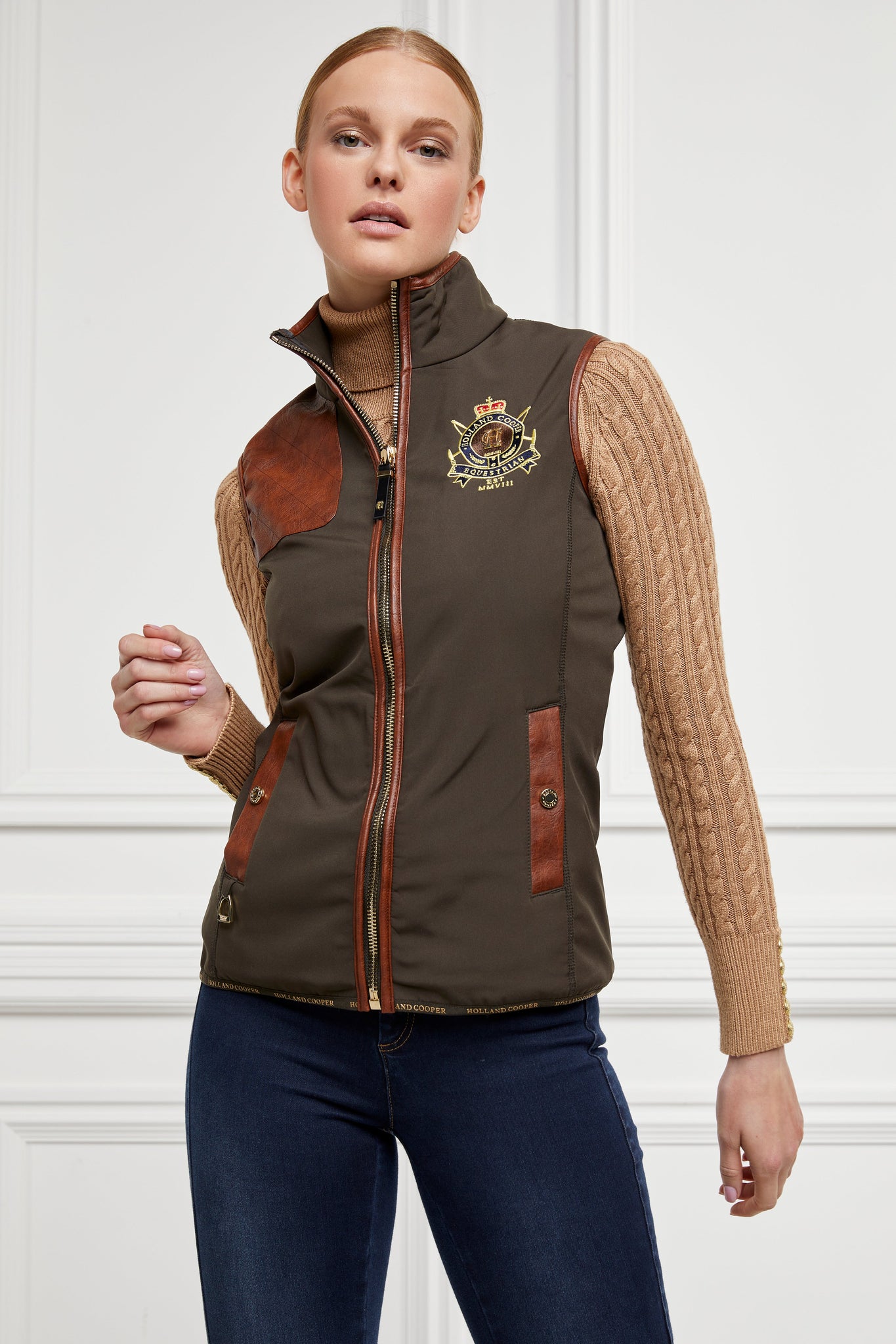 womens khaki gilet with dark brown leather seams along the arm holes pockets and down the zip with a gun patch on the shoulder and an embroidered logo worn with a beige knitted jumper and indigo jeans