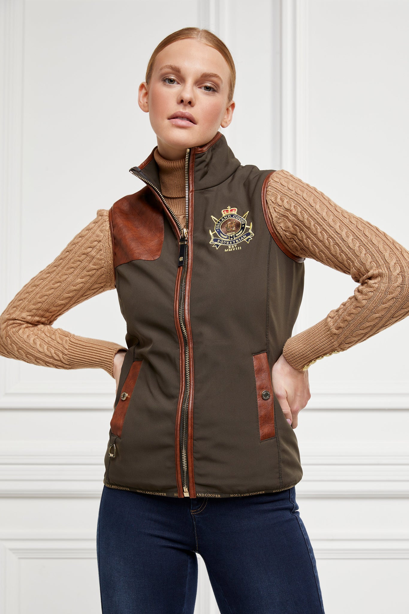 womens khaki gilet with dark brown leather seams along the arm holes pockets and down the zip with a gun patch on the shoulder  and an embroidered logo worn with a beige knitted jumper and indigo jeans