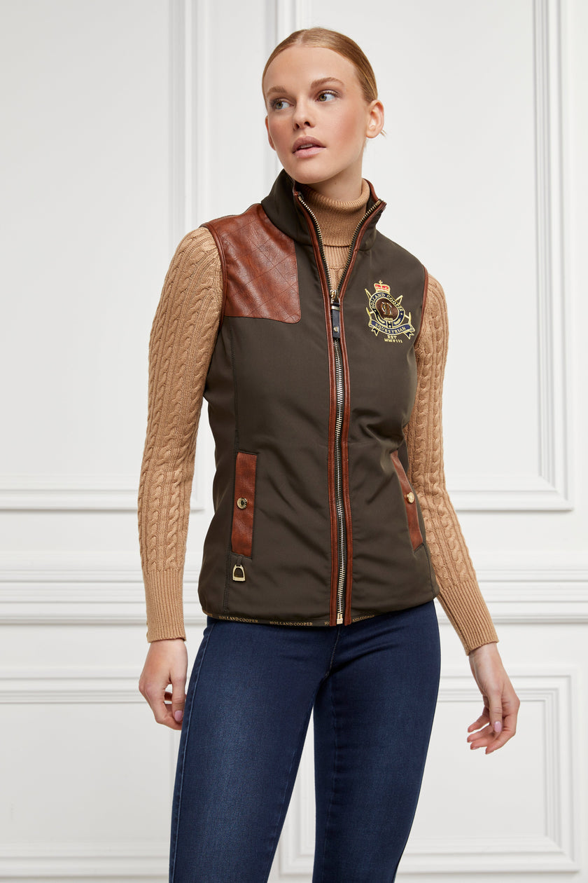 womens khaki gilet with dark brown leather seams along the arm holes pockets and down the zip with a gun patch on the shoulder and an embroidered logo worn with a beige knitted jumper and indigo jeans