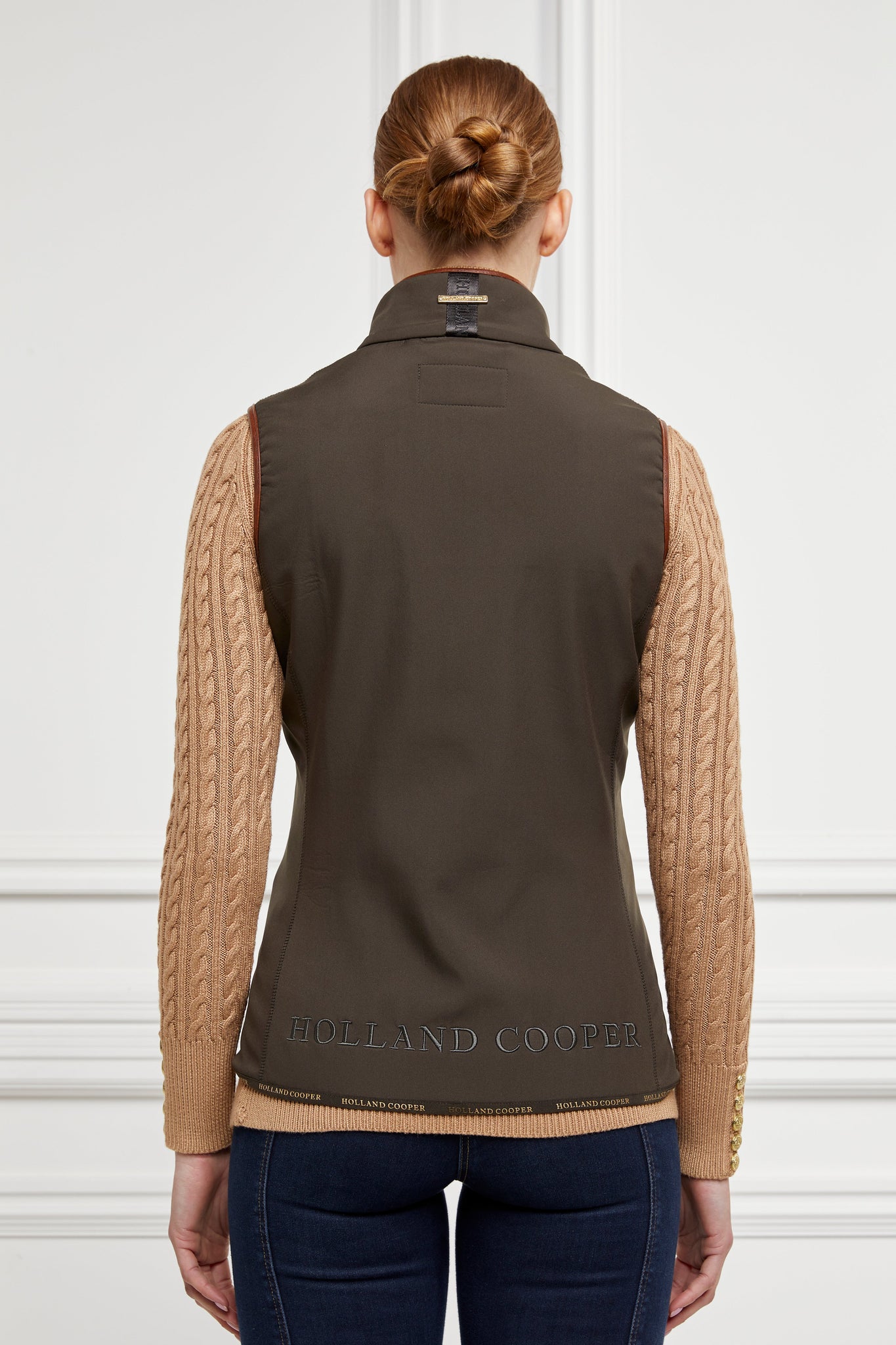 back of womens khaki gilet with dark brown leather seams and a logo across the bottom seam and on the collar