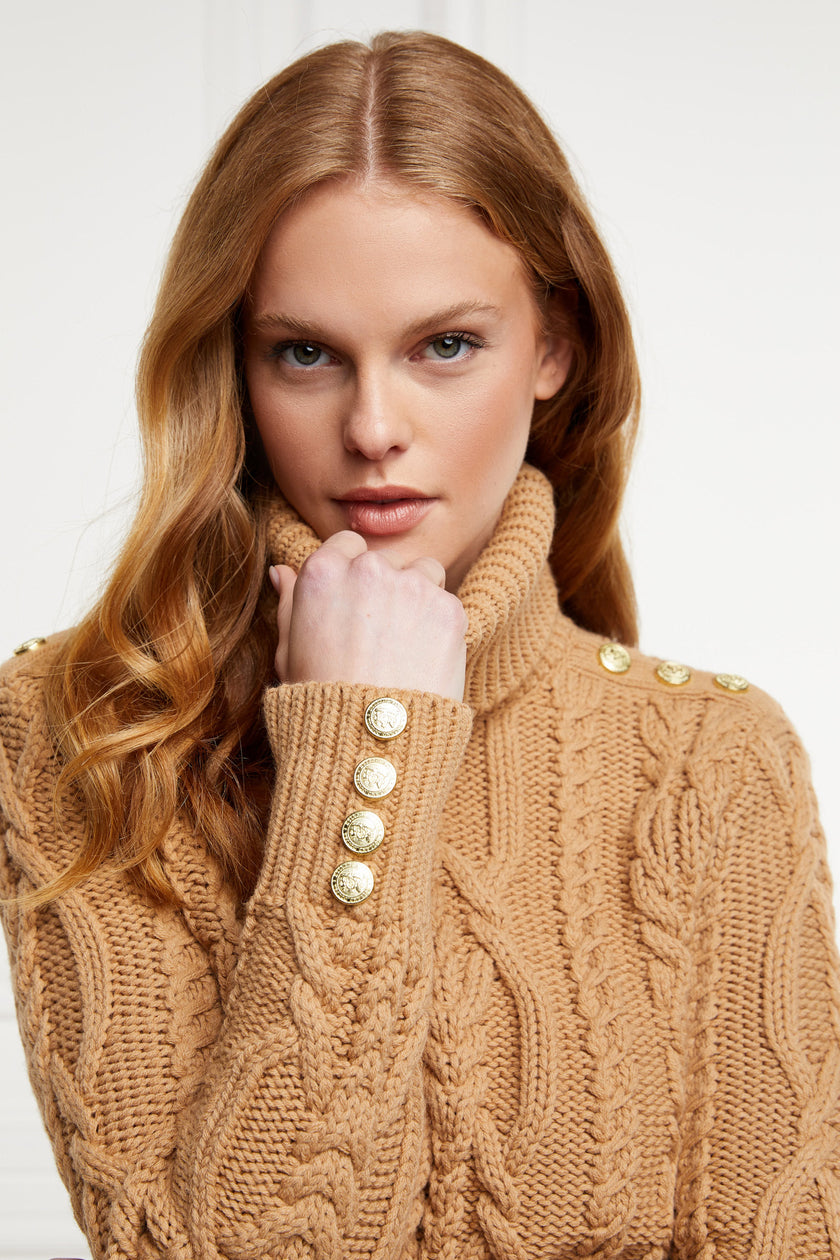 a chunky cable knit roll neck jumper in camel with dropped shoulders and thick ribbed cable trims and gold buttons on cuffs and collar