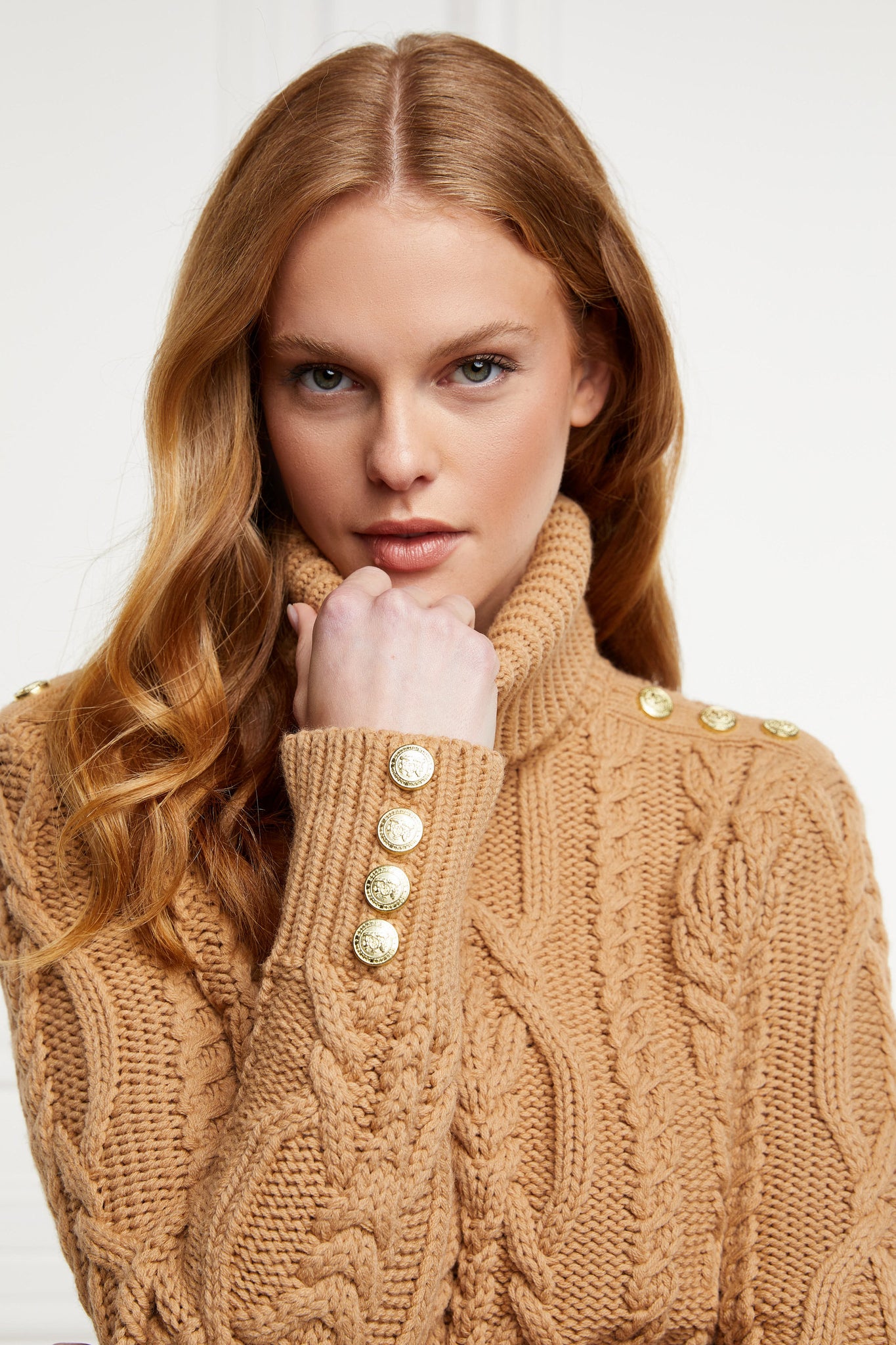 a chunky cable knit roll neck jumper in camel with dropped shoulders and thick ribbed cable trims and gold buttons on cuffs and collar