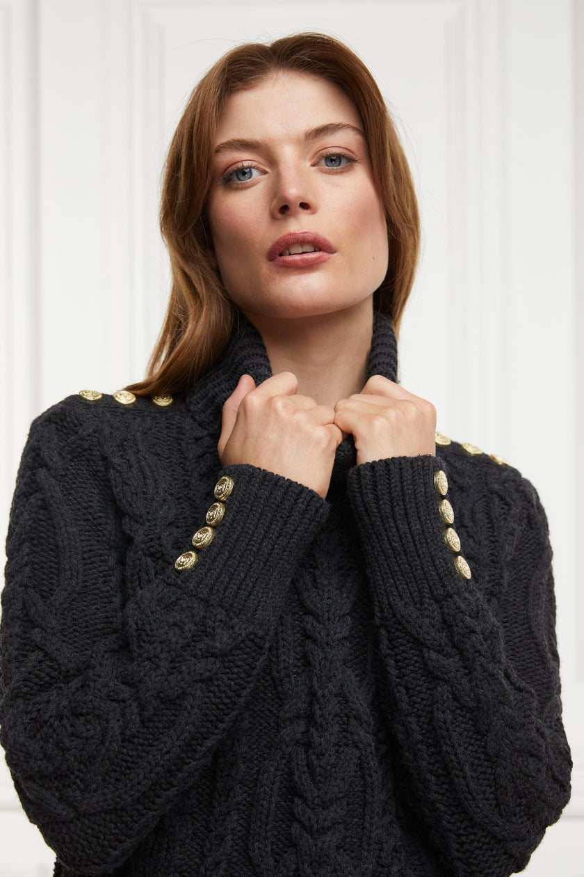 a chunky cable knit roll neck jumper in dark grey with dropped shoulders and thick ribbed cable trims and gold buttons on cuffs and collar