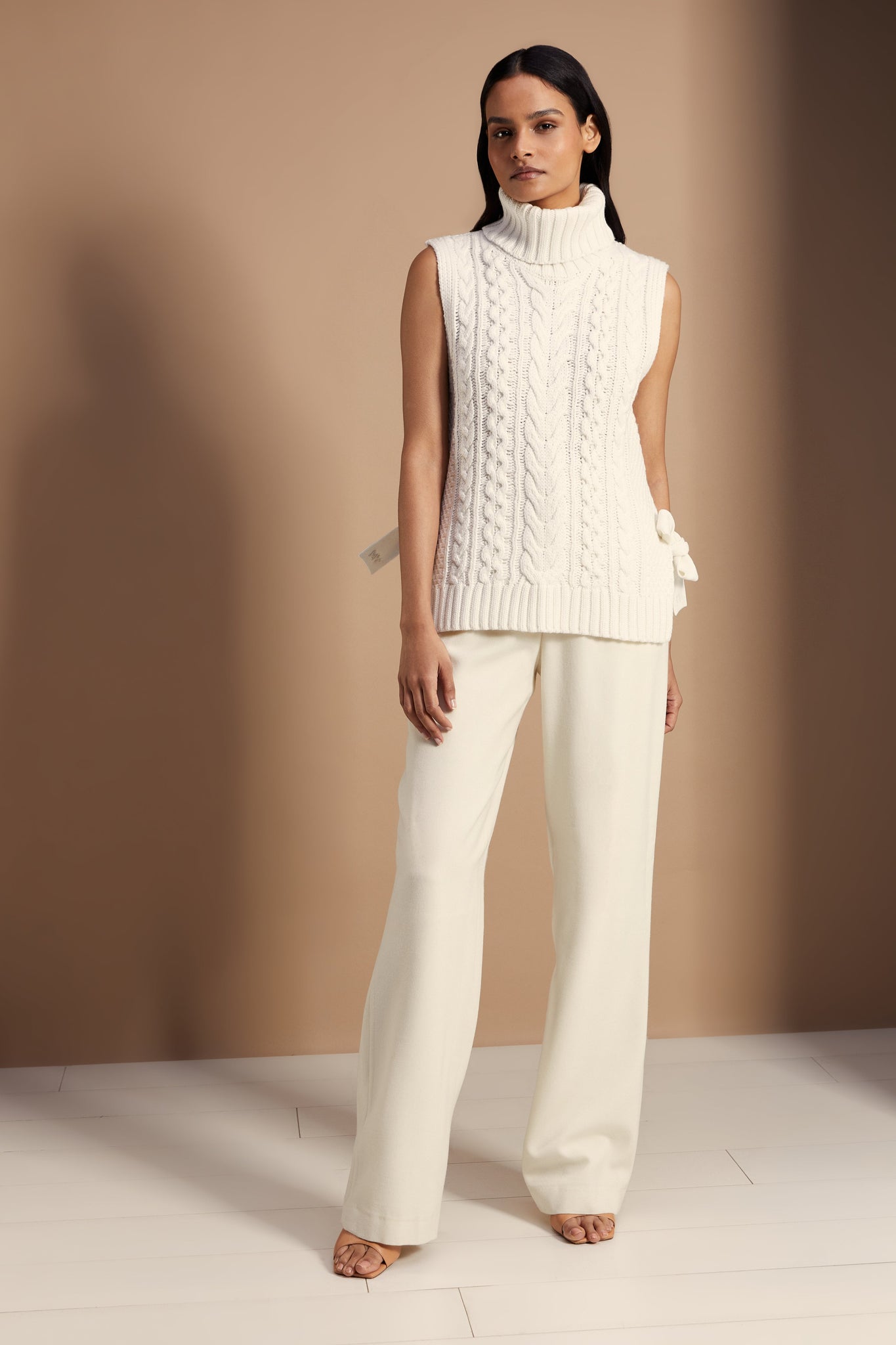 Sleeveless white chunky knit cable jumper with a roll neck and ties at each side 