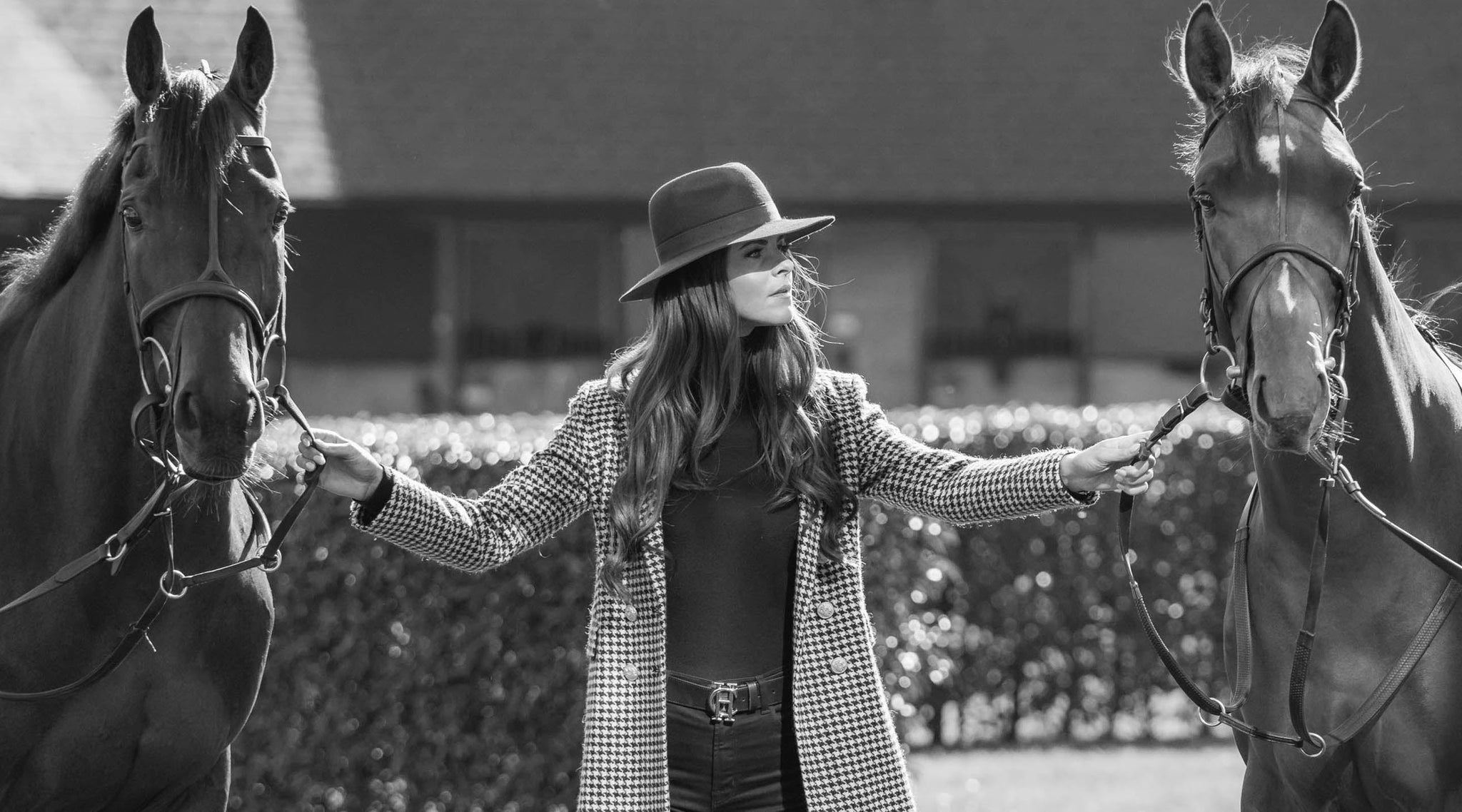 Black and white image with woman stood between two horses holding the bridle dressed in trilby hat and long houndstooth coat