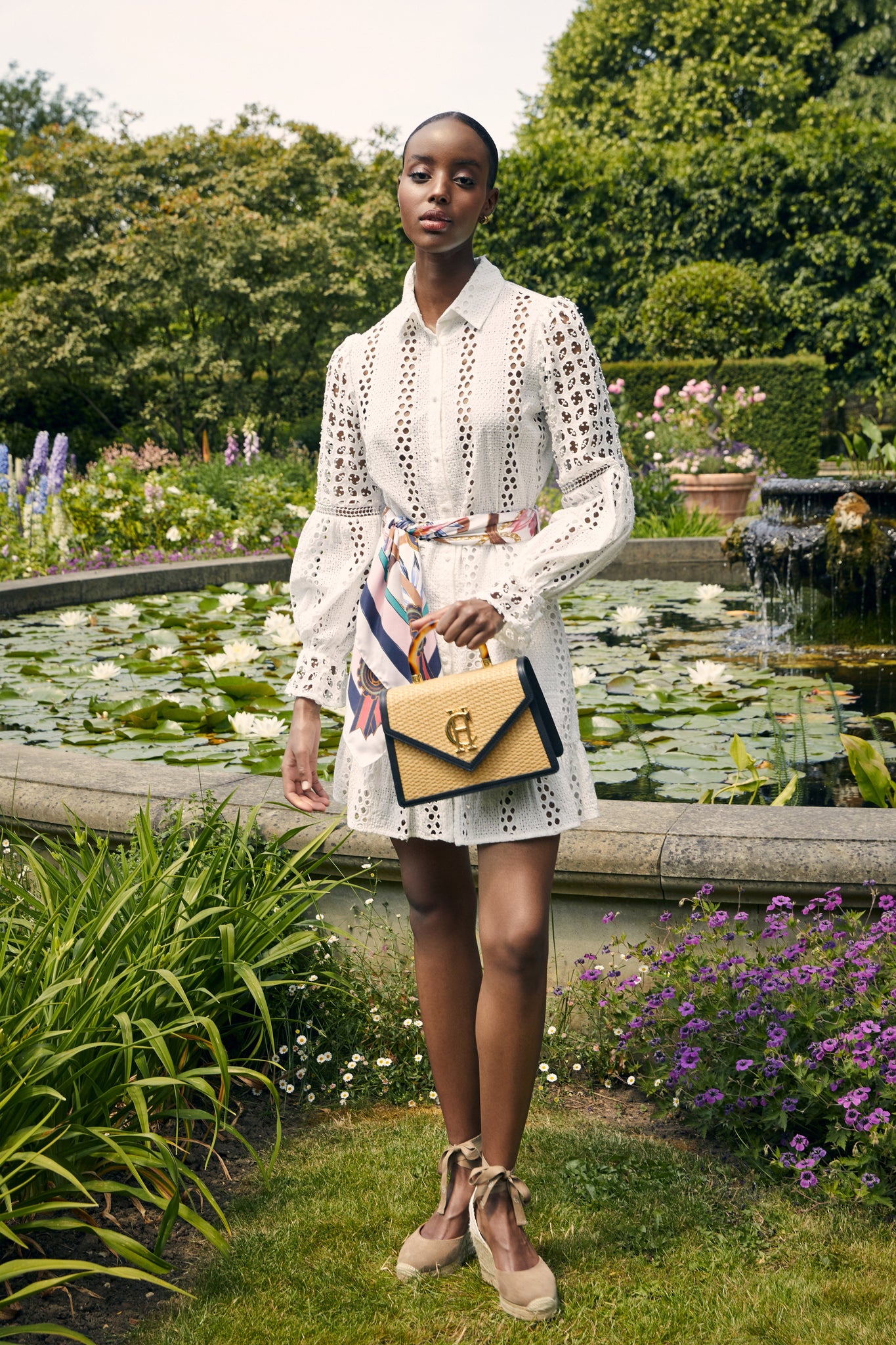 Womens white mini dress in broderie lace with a white and pink scarf around the waist and a black and natural raffia bag 