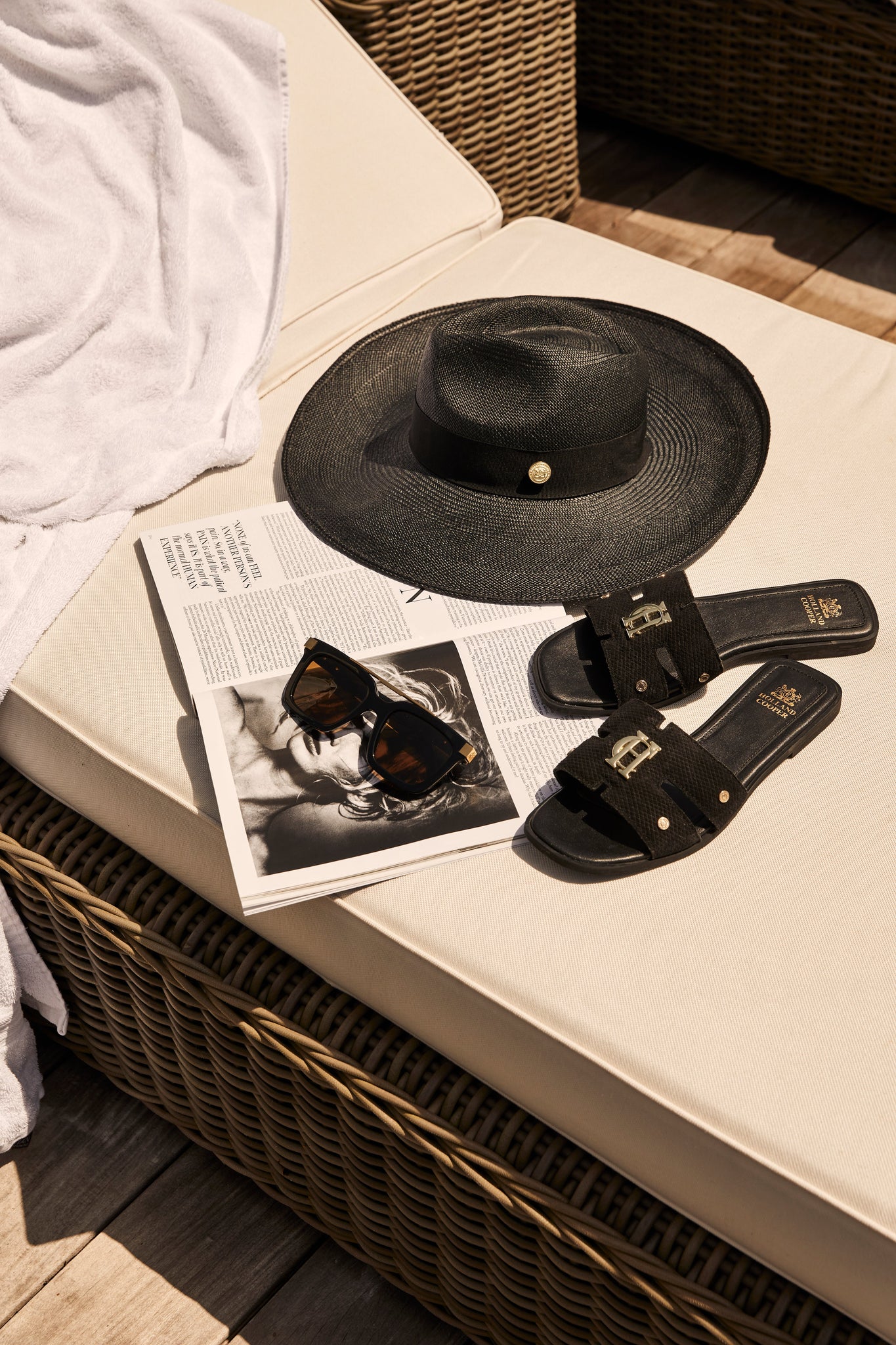 black suede sliders with a black leather sole and gold hardware sat on a sun bed next to black and gold sunglasses and a black straw hat