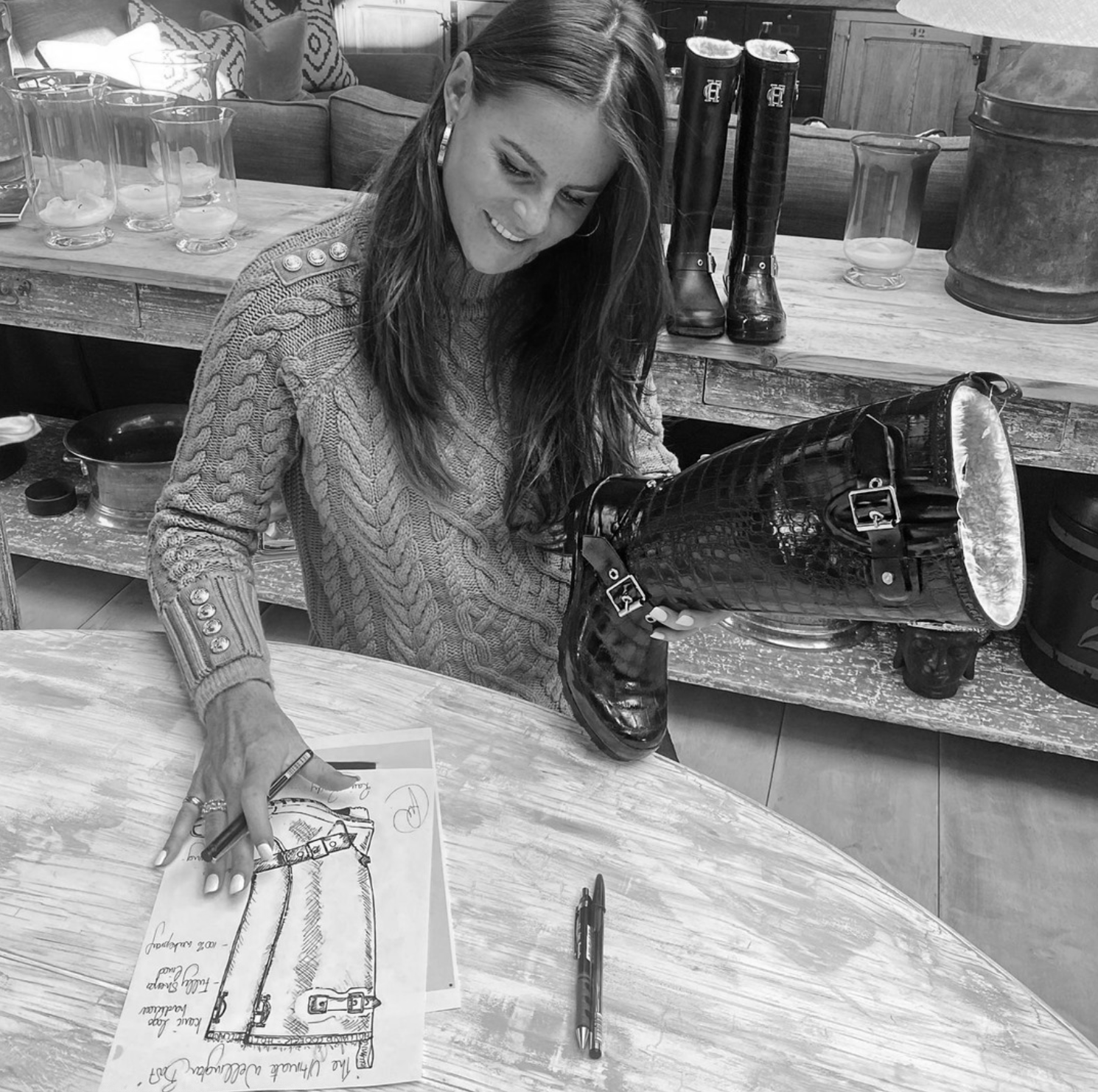 Black and white image of Jade Holland Cooper, owner of Holland Cooper, sat looking at sketches of wellingtons dressed in cable knit jumper holding pen and womens black croc wellington