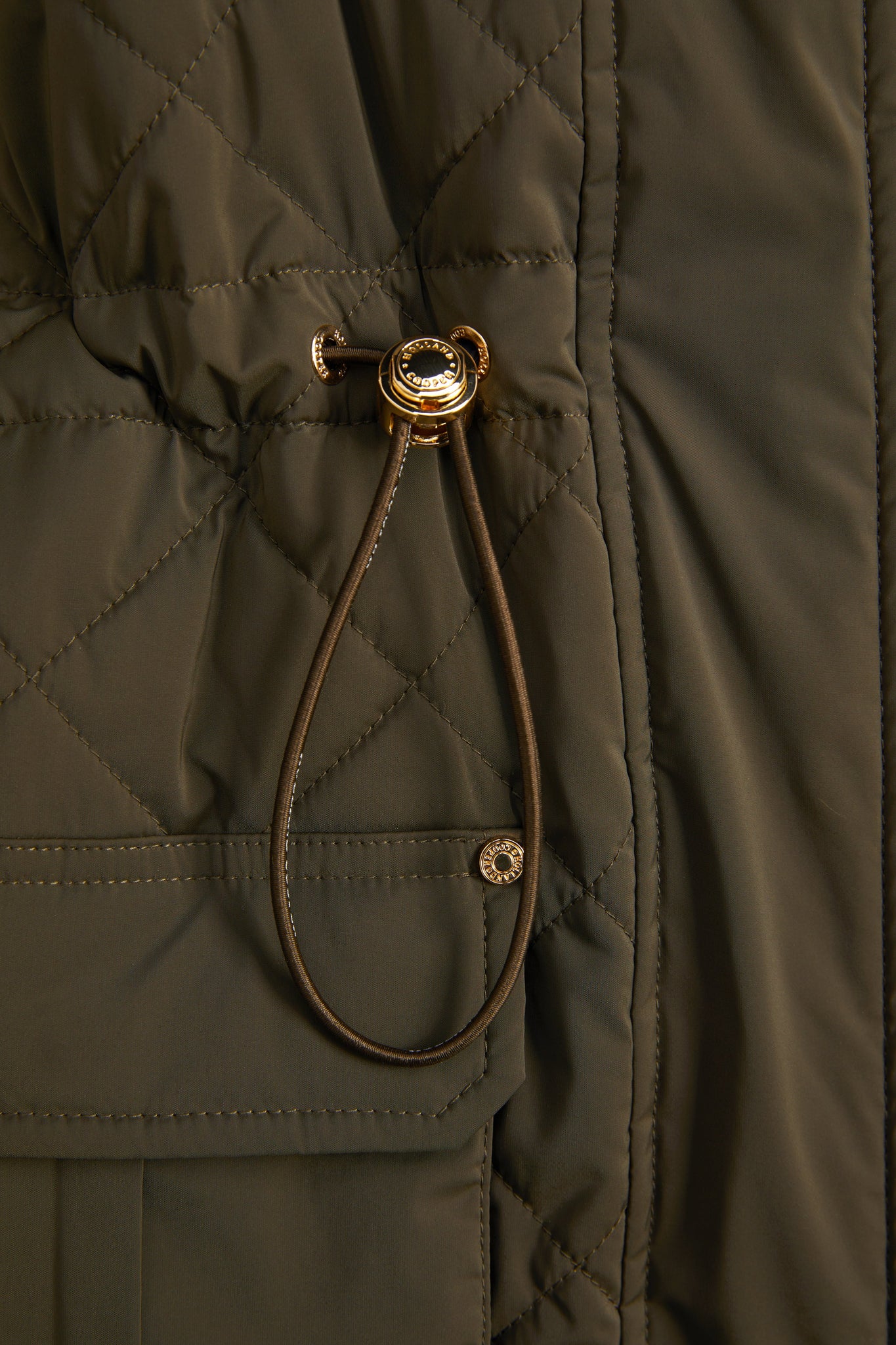 detail shot of the gold toggle on the womens khaki quilted gilet with a dark brown collar and seams along the arm holes with drawstring toggles on the waist and two front pockets