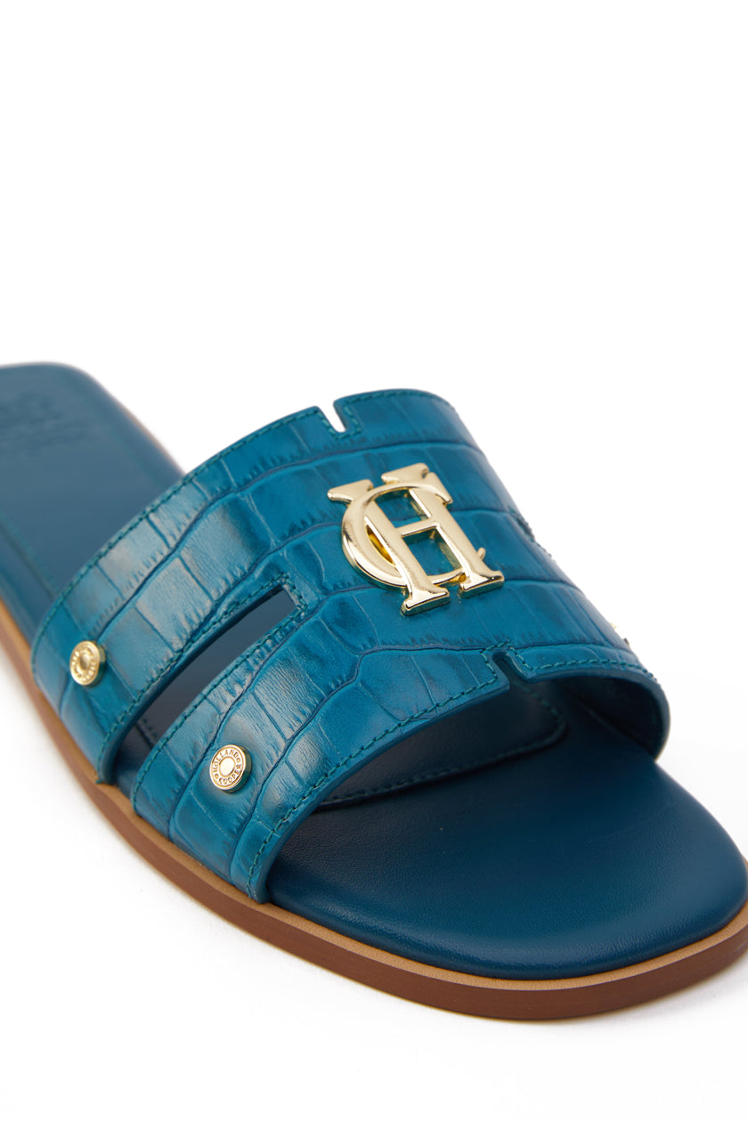 Close up of gold hardware on a teal blue croc embossed leather sliders with a tan leather sole