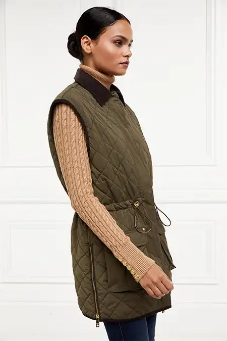 side of womens khaki quilted gilet with a dark brown collar and seams along the arm holes with drawstring toggles on the waist and two front pockets worn with a beige knitted jumper